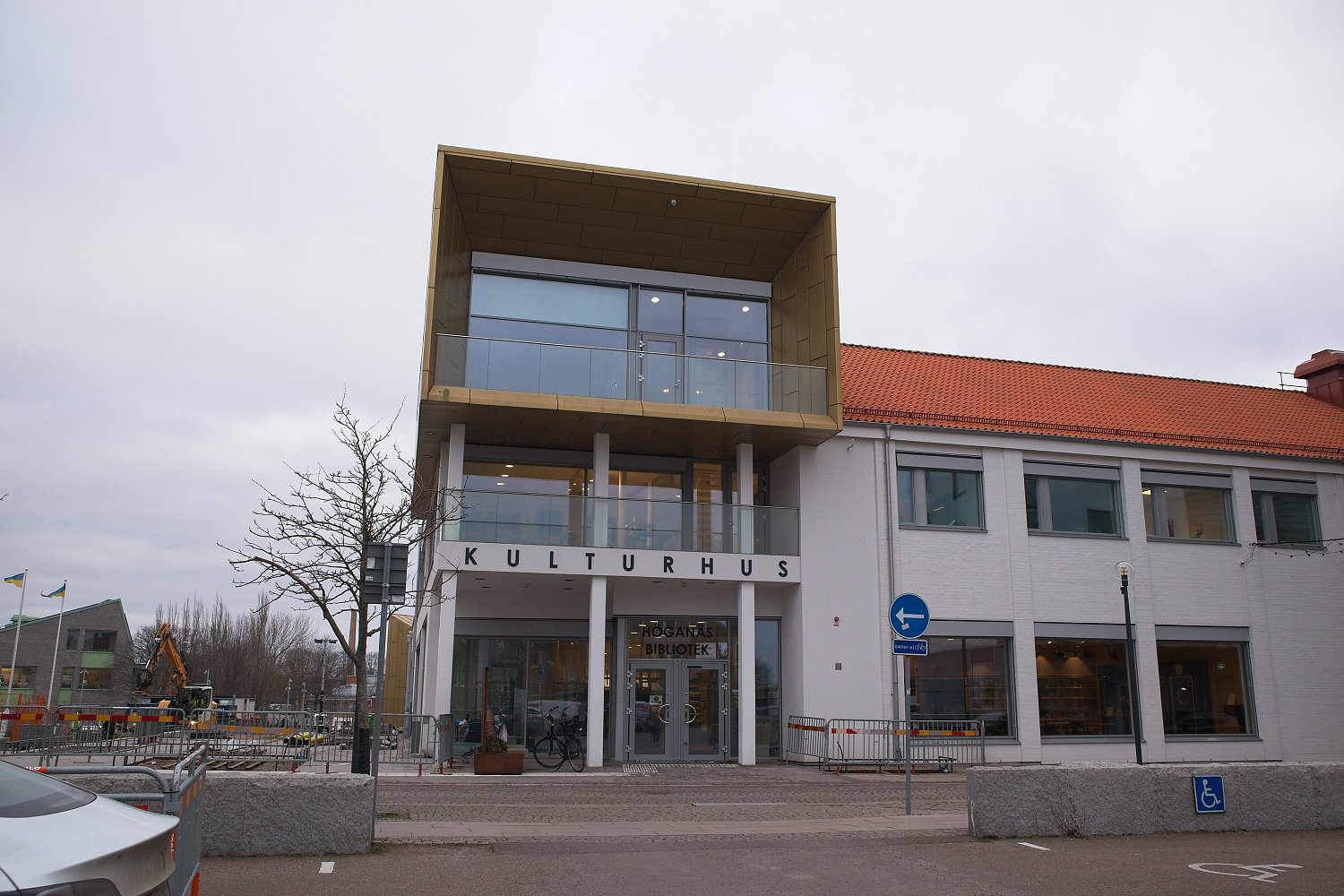 En fasadbild av en byggnad som heter KULTURHUS, med Höganäs Bibliotek vid entrén. Byggnaden har en modern del i guld och glas, samt en äldre vit del med rött tak. Framför byggnaden syns en parkeringsplats, en naken buske, ett cykelställ och inhägnat