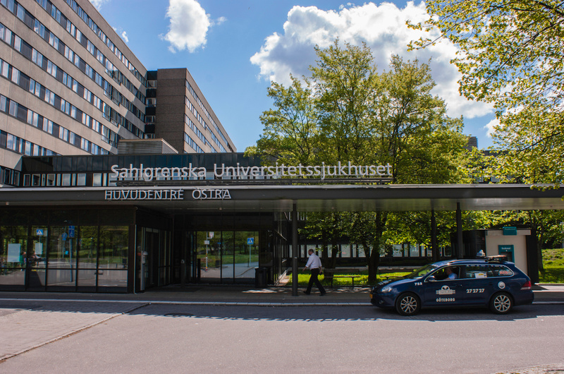 En bild som visar entrén till en byggnad märkt "Sahlgrenska Universitetssjukhuset HUVUDENTRÉ ÖSTRA" på en ljus dag. En mörk kombibil med texten "Göteborg" på sidan står parkerad nedanför en grön trädrad. En person går mot byggnaden till vänster.