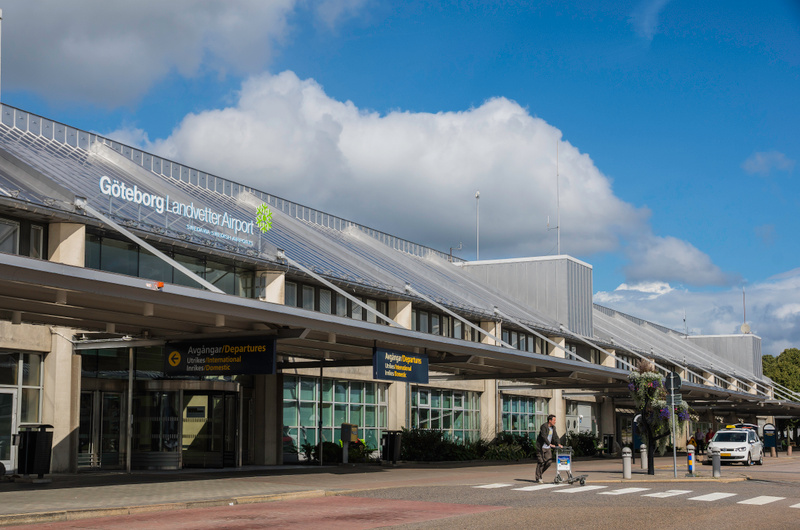 En fasadbild av Göteborg Landvetter Airport under dagtid med delvis molnig blå himmel. Byggnaden har ett glasat tak och en lång baldakin med skyltar för "Avgångar/Departures". En person med bagagevagn rör sig på gångvägen, och i högerkanten syns en v