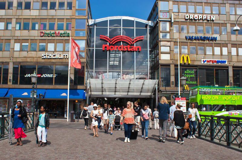 En folksamling går på plattlagd mark framför en stor byggnad med flera skyltar, bland annat "Nordstan" och "McDonald's". En grön "Hop On - Hop Off"-buss syns till höger.