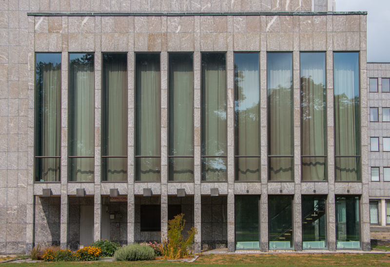 En fasadbild som visar en modern byggnad med en grå marmorerad yta och stora vertikala fönster. Fönstren är täckta av ljusa gardiner. Byggnaden har också mindre, fyrkantiga fönster på en sidofasad. Framför byggnaden finns gräs, buskar och blommor.