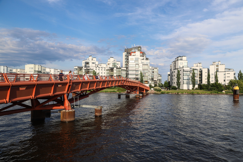 En röd bro sträcker sig över ett mörkt vatten under en klarblå himmel med moln. Längs flodkanten, på andra sidan bron, står modern arkitektur med vita byggnader. En gul boj syns i vattnet till höger om bron. Flera personer står på bron.