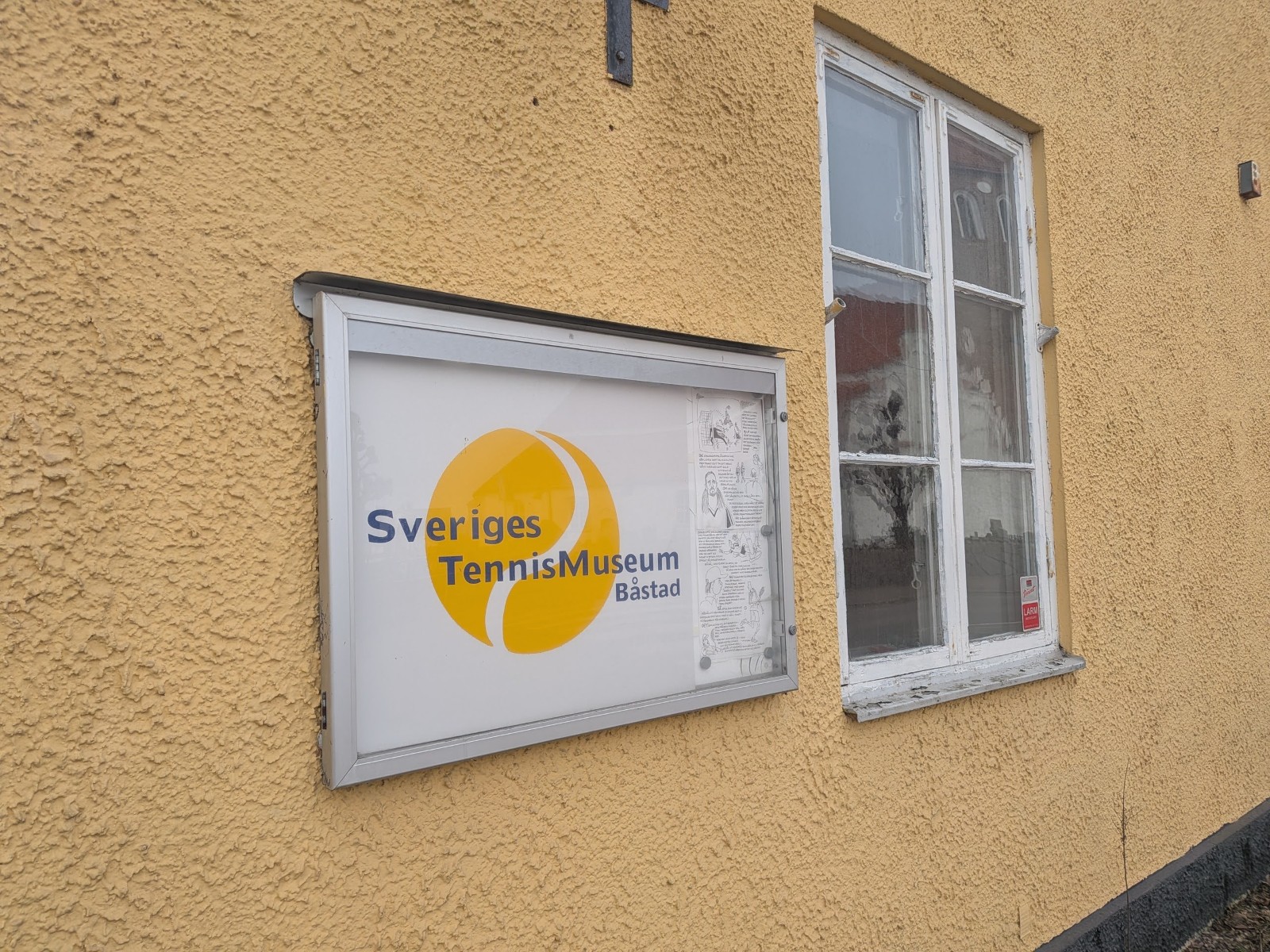 Bilden visar fasaden på en gul byggnad med en anslagstavla och ett fönster. Anslagstavlan har texten "Sveriges TennisMuseum Båstad" och en logotyp med en gul tennisboll. Fönstret intill har vita fönsterkarmar och reflekterar omgivningen.