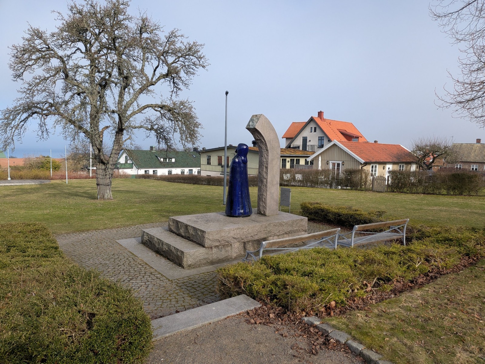 En bild som visar en parkmiljö med en central skulptur, ett stort träd utan löv, gräsmattor och buskar. I bakgrunden syns flera hus med röda och gröna tak. En grusgång och stentrappa leder till skulpturen.