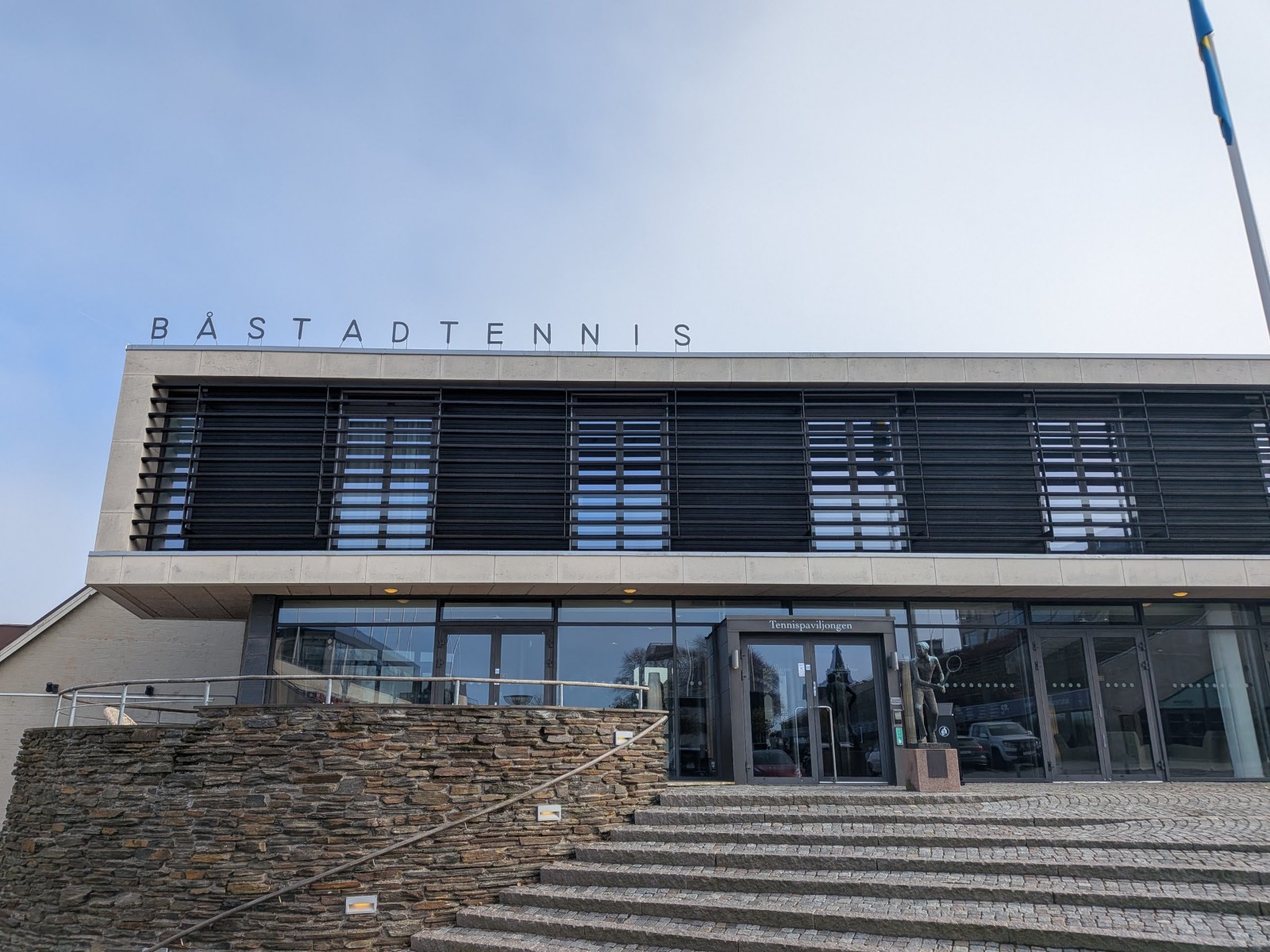 En modern byggnad delvis klädd i sten med stora fönster och en skylt där det står "BÅSTADTENNIS" på taket. Trappor av gatsten leder upp till en entré där det även står "Tennispaviljongen". Bredvid entrén står en staty och en flaggstång med en blå fla