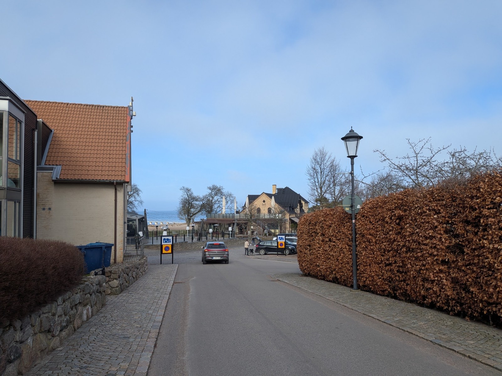 En gata leder mot en anläggning med bebyggelse, strand och hav i bakgrunden. En bil kör bort från kameran mot den anläggningen. Längs vänster sida syns en del av en byggnad med tegeltak och en stenmur. På höger sida finns en hög, brun häck och en gat