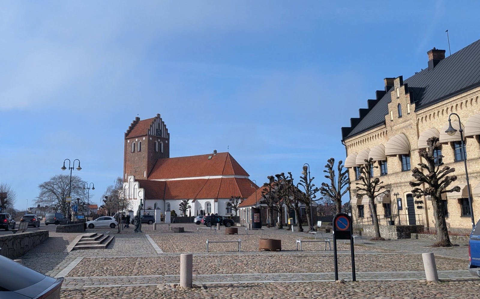 En stor stenbelagd öppen plats med en äldre röd tegelkyrka och en annan större gul tegelbyggnad med mörkt tak. På platsen finns träd utan löv, lyktstolpar och bänkar. Till vänster passerar bilar och människor syns på avstånd.