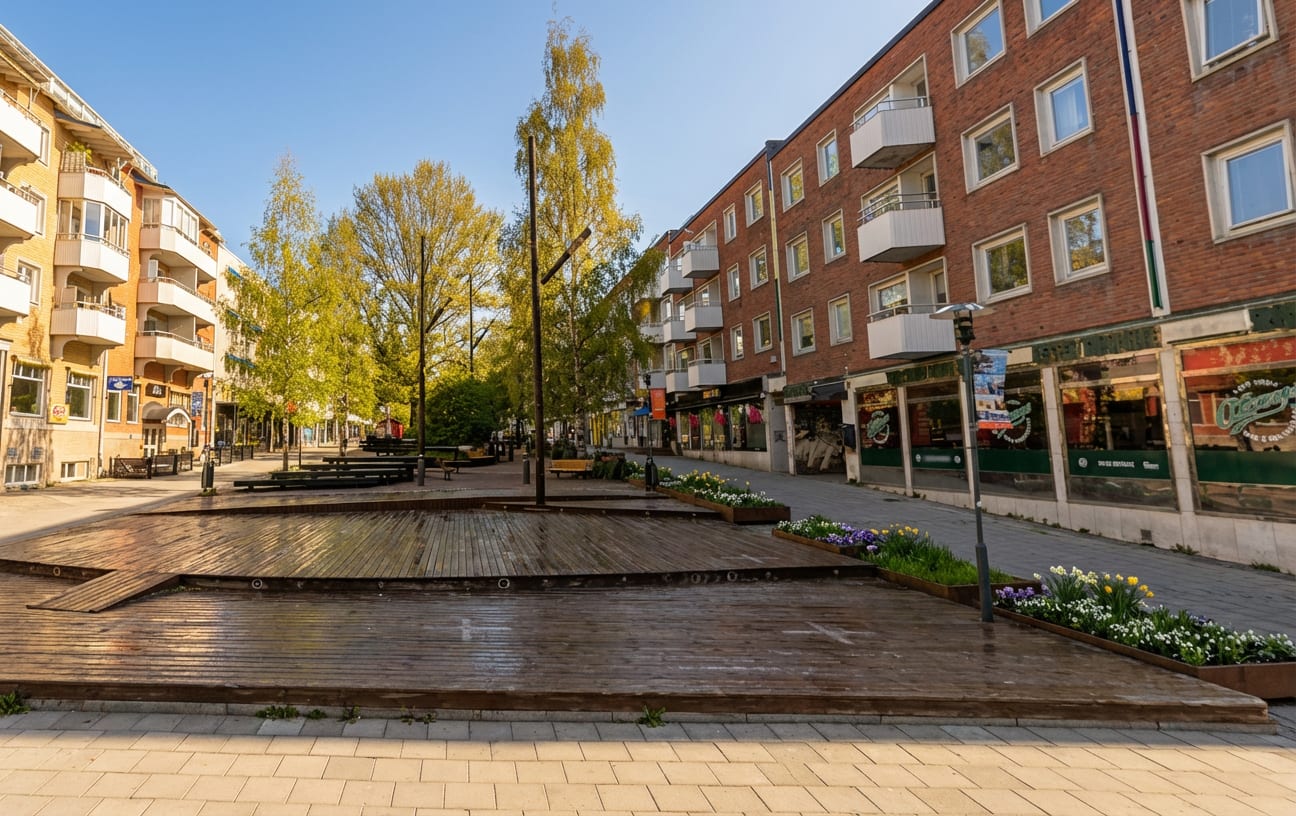 En bild som visar en gågata med en stor träplattform i förgrunden. Längs gatan med grönskande träd är byggnader med butiksfasader och balkonger. På träplattformen och i blomlådor finns växter och blommor.