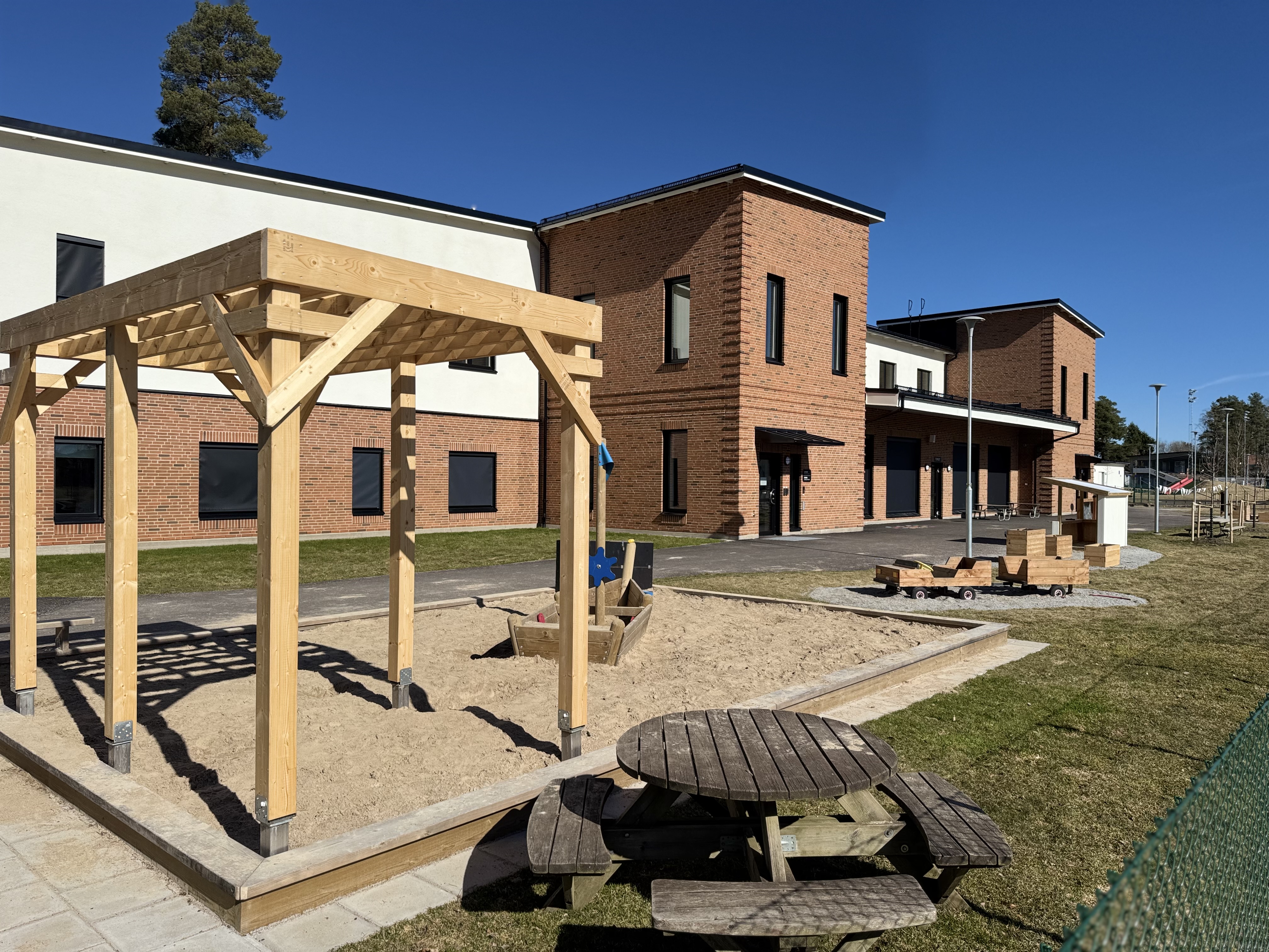 En utomhusbild föreställande en skola eller förskola med en stor sandlåda i förgrunden. Till vänster i sandlådan finns en träkonstruktion med tak, och i mitten ett träfartyg. En träbänk med bord samt trävagnar syns också. Byggnader med tegelfasader o