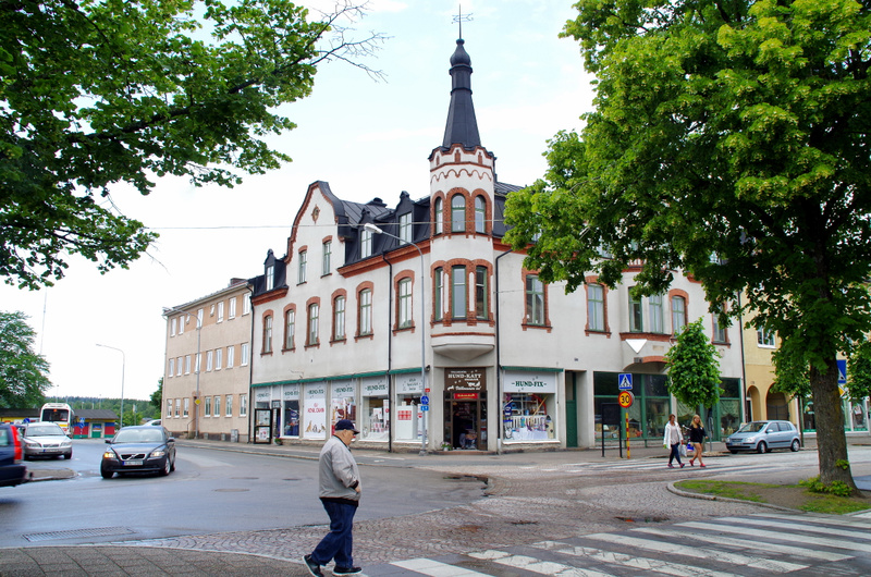 Bilden visar gatukorsningen framför en större vit byggnad med ett torn och flera butiker på bottenplan. En person går över ett övergångsställe i förgrunden. I bakgrunden syns bilar och träd med gröna löv.