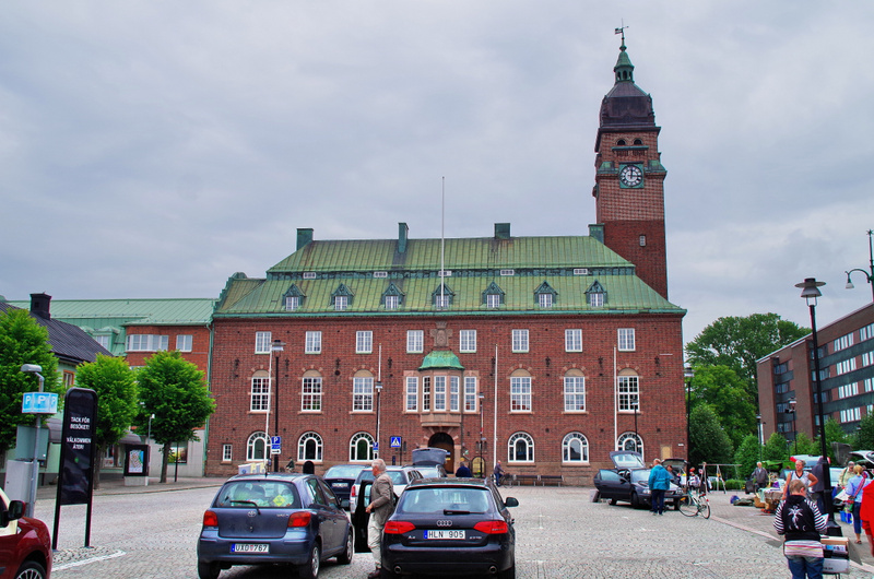 En stor, röd tegelbyggnad med grönt tak och ett klocktorn dominerar bilden. Framför byggnaden finns en kullerstenstorg med parkerade bilar och människor. På torget syns även en gatumarknad med stånd och besökare.