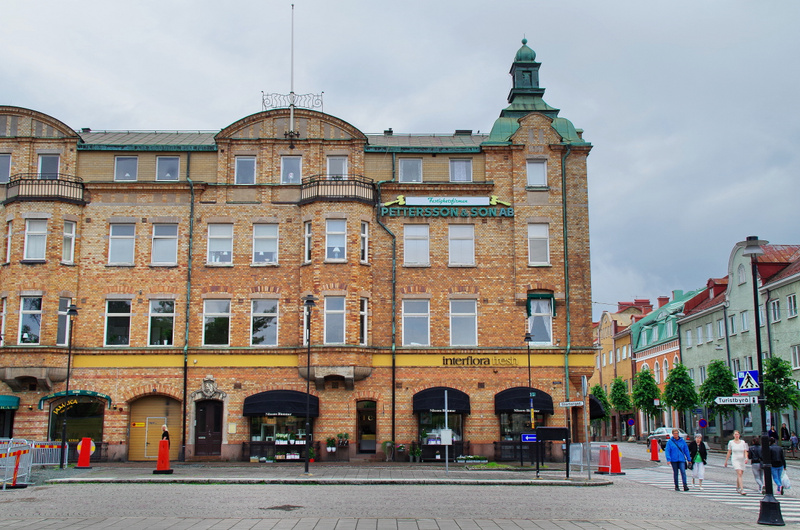 En stor, tegelröd byggnad i flera våningar med vita fönster, balkonger och ett grönt torn. På fasaden finns skyltar för "PETTERSSON & SON AB" och "interflora fresh". Gator syns med trafikkoner, gatuskyltar och en zebraövergång där flera personer går.