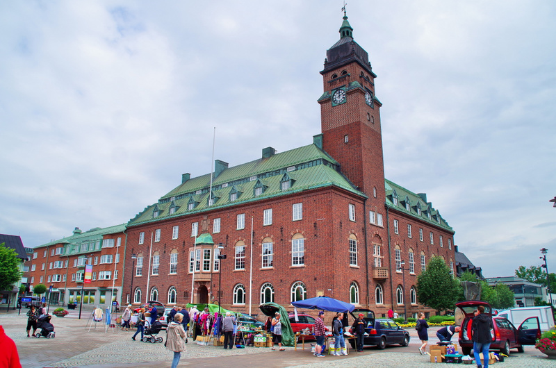En stor, röd tegelbyggnad med en klocktorn och grönt tak dominerar bilden. Framför byggnaden pågår en marknad eller loppis med flera stånd och människor som tittar på varor. Himmel är mulen.