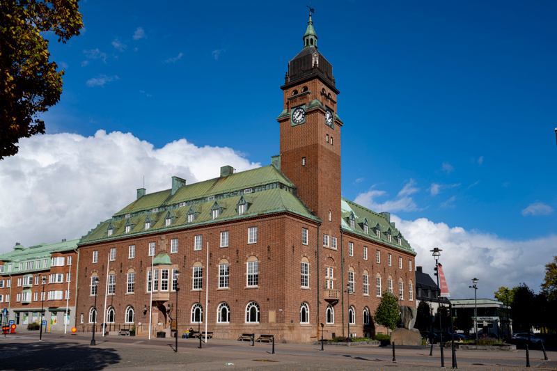 En stor, röd tegelbyggnad med ett högt klocktorn och ett grönt tak dominerar bilden. Byggnaden har många fönster och balkonger. I förgrunden syns en öppen plats med gatlyktor, några bänkar samt träd och buskar. Himlen är blå med vita moln.
