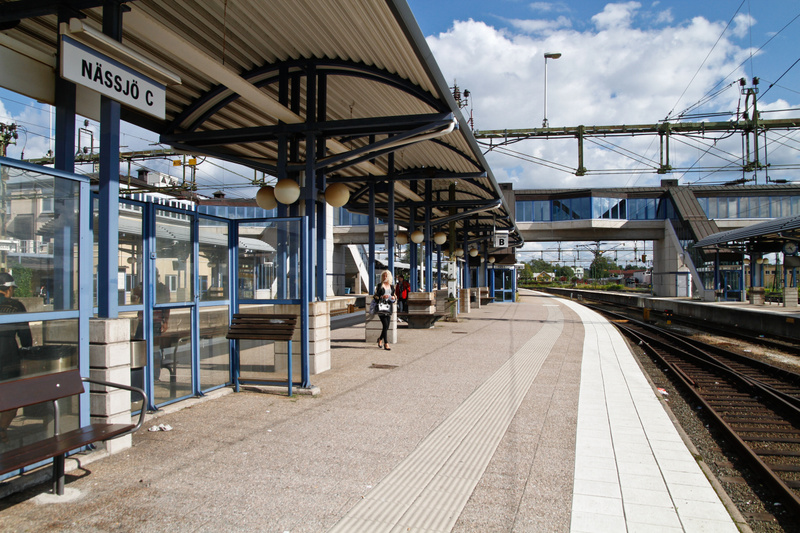 En bild av en tågperrong under dagtid med en kvinna gående och en skylt som indikerar 'NÄSSJÖ C'. Perrongen har skärmtak, bänkar och ett tak som sträcker sig över delar av perrongen. I bakgrunden syns tågspår, en gångbro och en klarblå himmel med mol