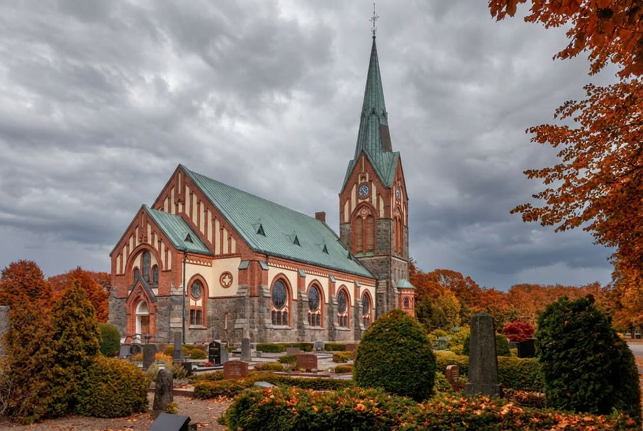 En kyrka med tegel- och stenfasad samt grön tak reser sig mot en molnig himmel. Runt kyrkan finns en kyrkogård med gravstenar och höstfärgade träd och buskar. Löv i höstfärger syns i förgrunden och bakgrunden.