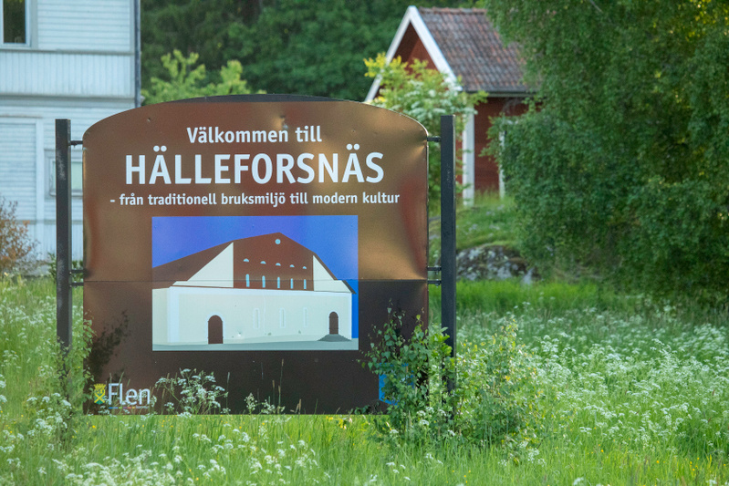 En stor brun skylt med texten "Välkommen till HÄLLEFORSNÄS - från traditionell bruksmiljö till modern kultur" och en illustration av en byggnad står i en grön gräsyta med vita blommor. I bakgrunden syns skymten av byggnader och grönskande träd och bu