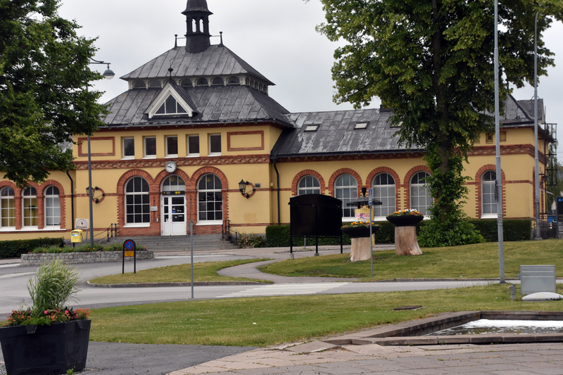 En gul byggnad med bruna tegeldetaljer och ett mörkt tak. Utanför byggnaden finns ett gräsområde, träd, en gångväg och en väg. På gräsområdet står två blomkrukor på trästammar och en svart informationstavla. En gul brevlåda och en cykelparkeringsskyl