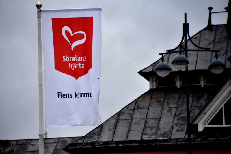 En vit flagga med texten "Sörmlands hjärta" och "Flens kommun" samt en röd sköld med ett vitt hjärta, syns mot en grå himmel. Till höger syns delar av ett mörkt tak med dekorativa element och en gatlykta med tre runda lampor.