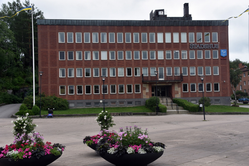 En fasadvy av en röd tegelsbyggnad med skylten "STADSHUS" ovanför ingången. Framför byggnaden finns ett öppet torg med större blomkärl fyllda med växter i olika färger, samt en flaggstång med en vajande svensk flagga. I bakgrunden syns skog och en ku