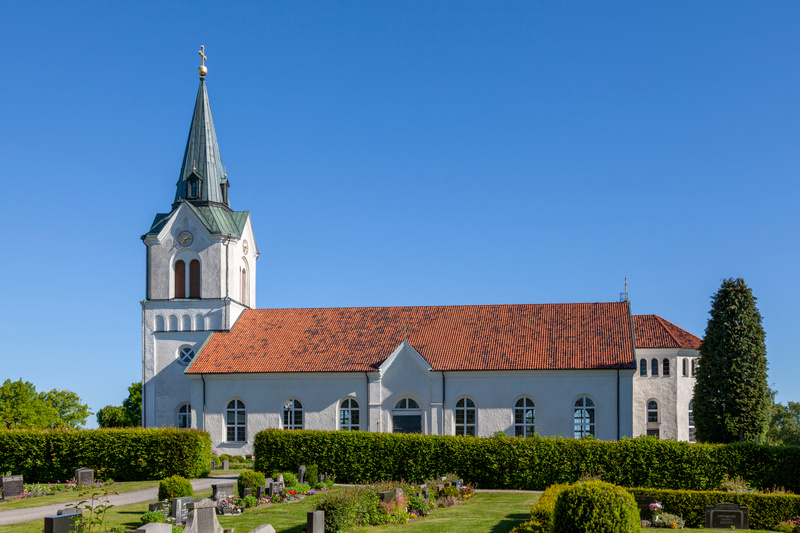 En vit kyrka med rött tak och en hög grön spira syns under en molnfri blå himmel. Framför kyrkan finns en grön häck och en kyrkogård med gravstenar och gräsmattor.