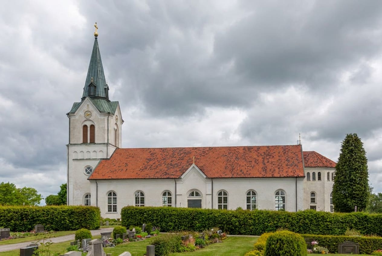 En vit kyrka med rött tak och en grön spira syns under en molnig himmel. Framför kyrkan finns en grön gräsmatta, buskar och gravstenar. Spiran har ett gyllene kors på toppen och en urtavla på kyrktornet.