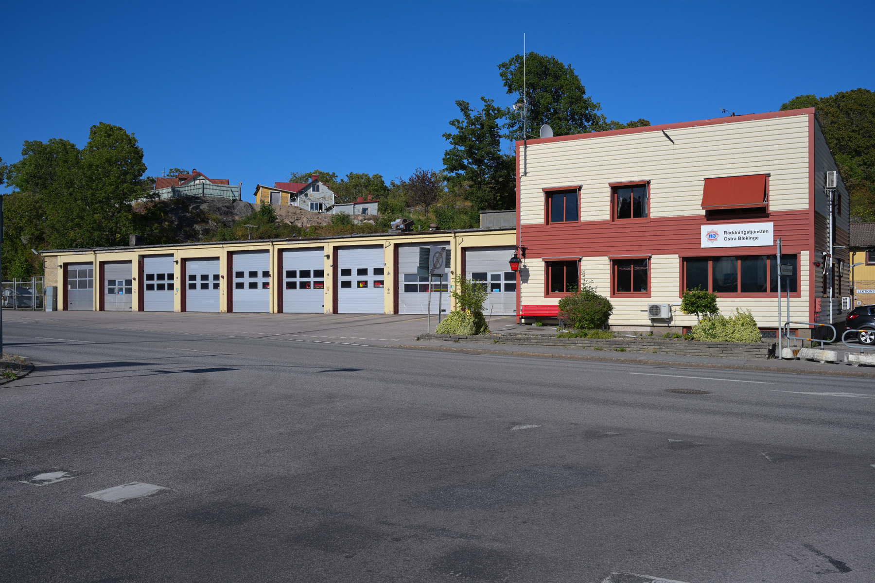 En rymlig utomhusbild som visar en brandstation med flera garageportar en solig dag. Till höger finns en tvåvåningsbyggnad med en skylt som indikerar 'Räddningstjänsten Östra Blekinge'.