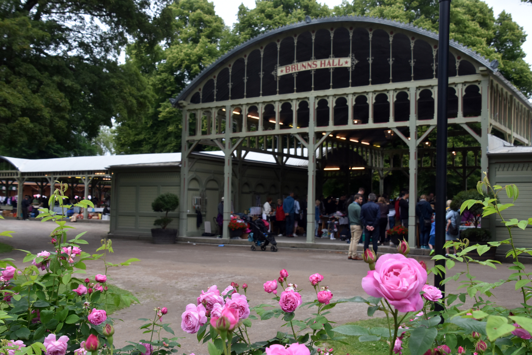 En bild visar en utomhusmiljö med en byggnad som har en skylt med texten "BRUNNS HALL". På bilden syns flera människor samlade, samt en perambulator. I förgrunden blommar rosa rosor. Bakom byggnaden finns träd med gröna löv.