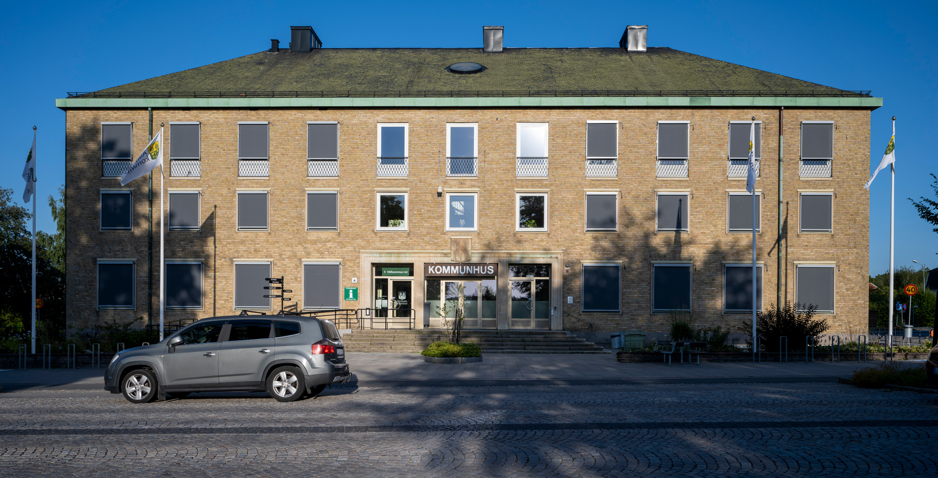 En gul tegelbyggnad med texten "KOMMUNHUS" ovanför entrén syns i solljus, med en grå bil parkerad framför. Byggnaden har flera fönster, med mörka jalusier på många av dem. Två flaggstänger med vita flaggor står på var sida om byggnaden.