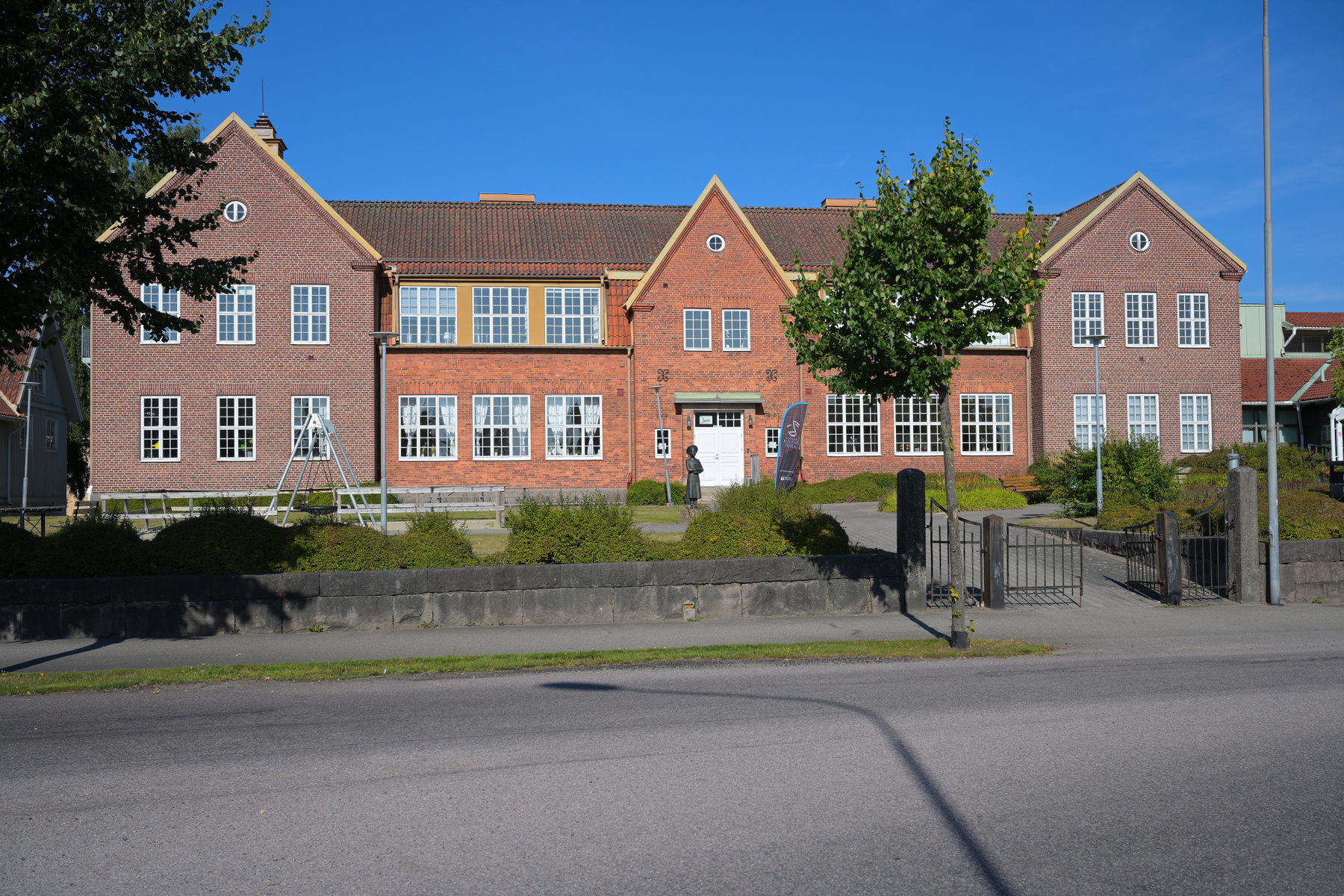 En stor, röd tegelbyggnad med vita fönsterkarmar och ett brunt tegeltak. Framför byggnaden syns en asfalterad väg, en stenmur, gräs och buskar. En svart staty av en liten person står framför huvudentrén, och till höger om den finns en blå flagga.