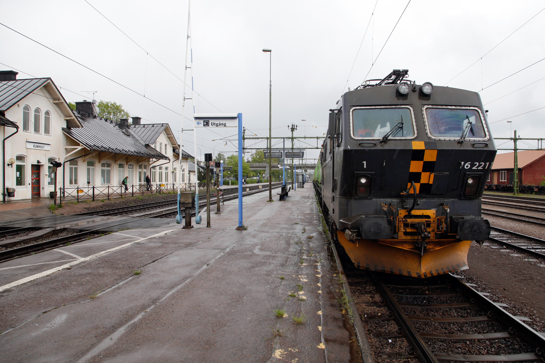 En bild som visar ett tåg vid en perrong på en järnvägsstation under en molnig dag. På vänster sida syns stationsbyggnader i ljus färg med svarta tak. Ett stort lok med nummer 162211 står stilla vid perrongen. Skylten "ÄLMHULT" syns