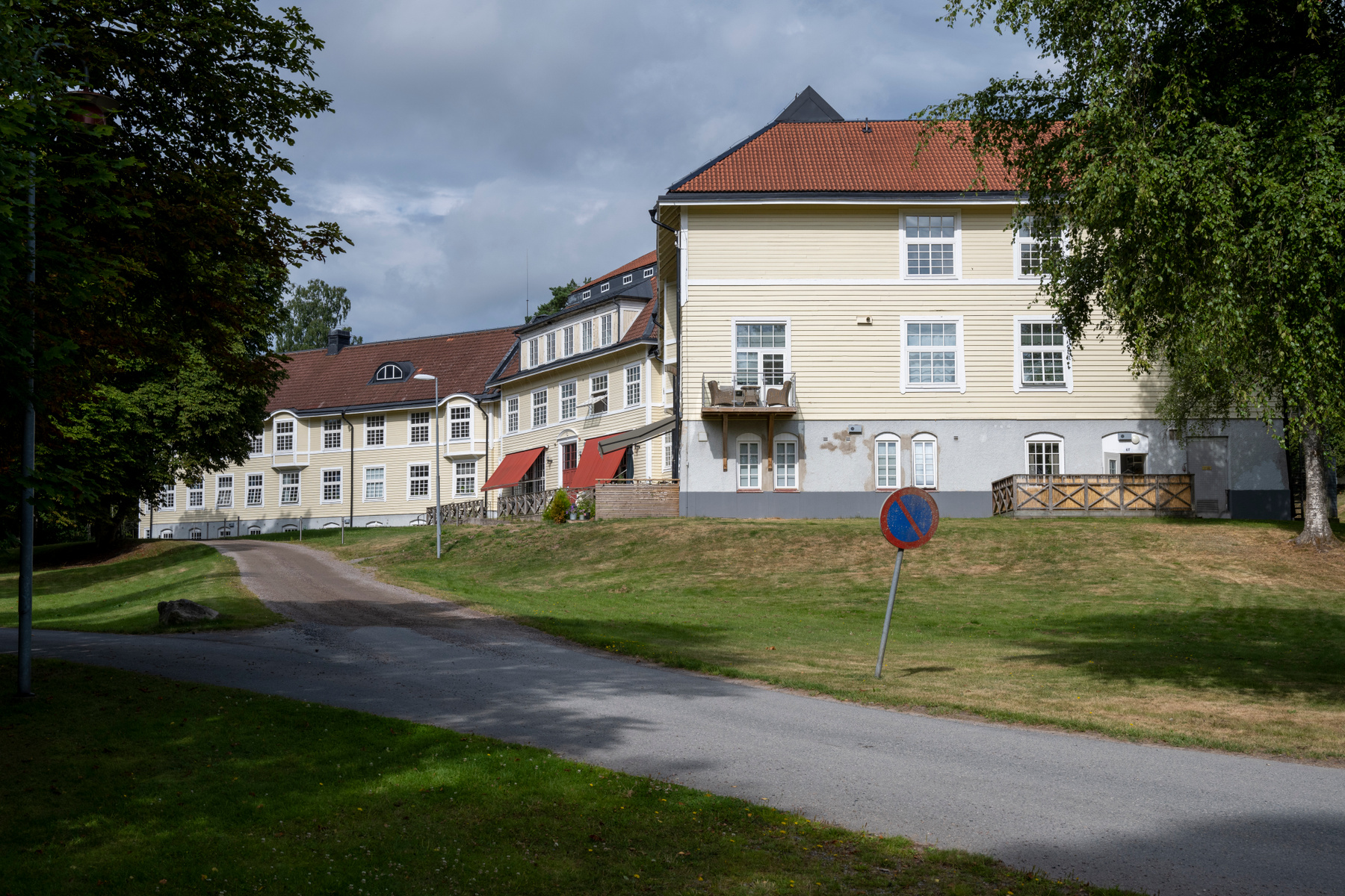 En gul byggnad med rött tak och verandor, omgiven av gräsmattor och träd under en molnig himmel. Framför byggnaden finns en asfalterad väg och en skylt som indikerar stoppförbud. Till vänster och höger om byggnaden syns gröna lövträd.