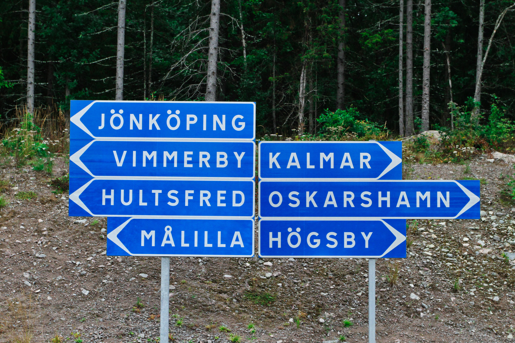 En bild som visar en blå vägskylt med vita textfält som pekar mot olika svenska orter, bland annat Jönköping, Vimmerby, Kalmar, Hultsfred, Oskarshamn, Målilla och Högsby. Bakgrunden består av en grön skog och grus. Bilden är tagen utomhus.