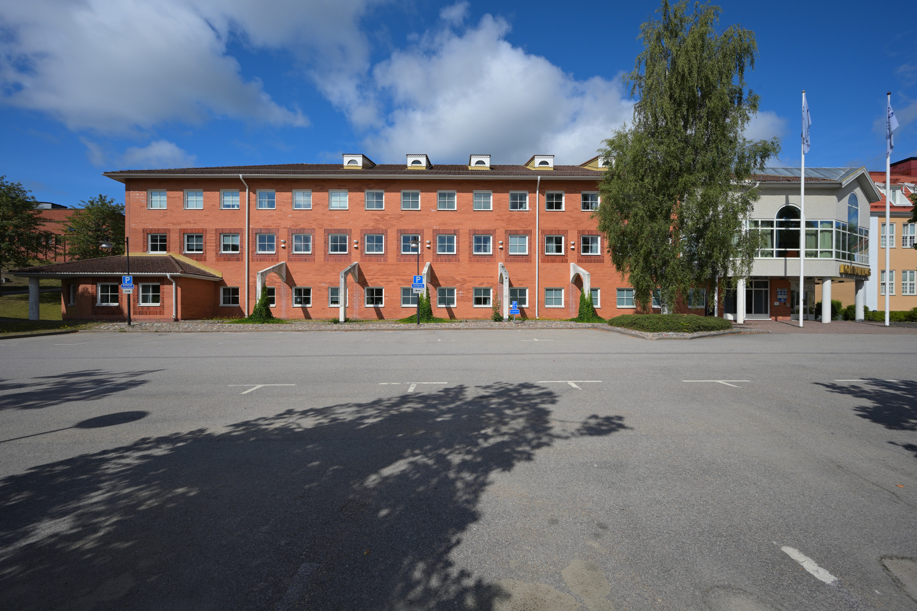 En stor, röd tegelbyggnad med många fönster står under en blå himmel med vita moln, framför en asfalterad parkeringsplats där skuggor från träd faller. Ett parkeringsskylt syns tydligt på vänster sida av bilden.