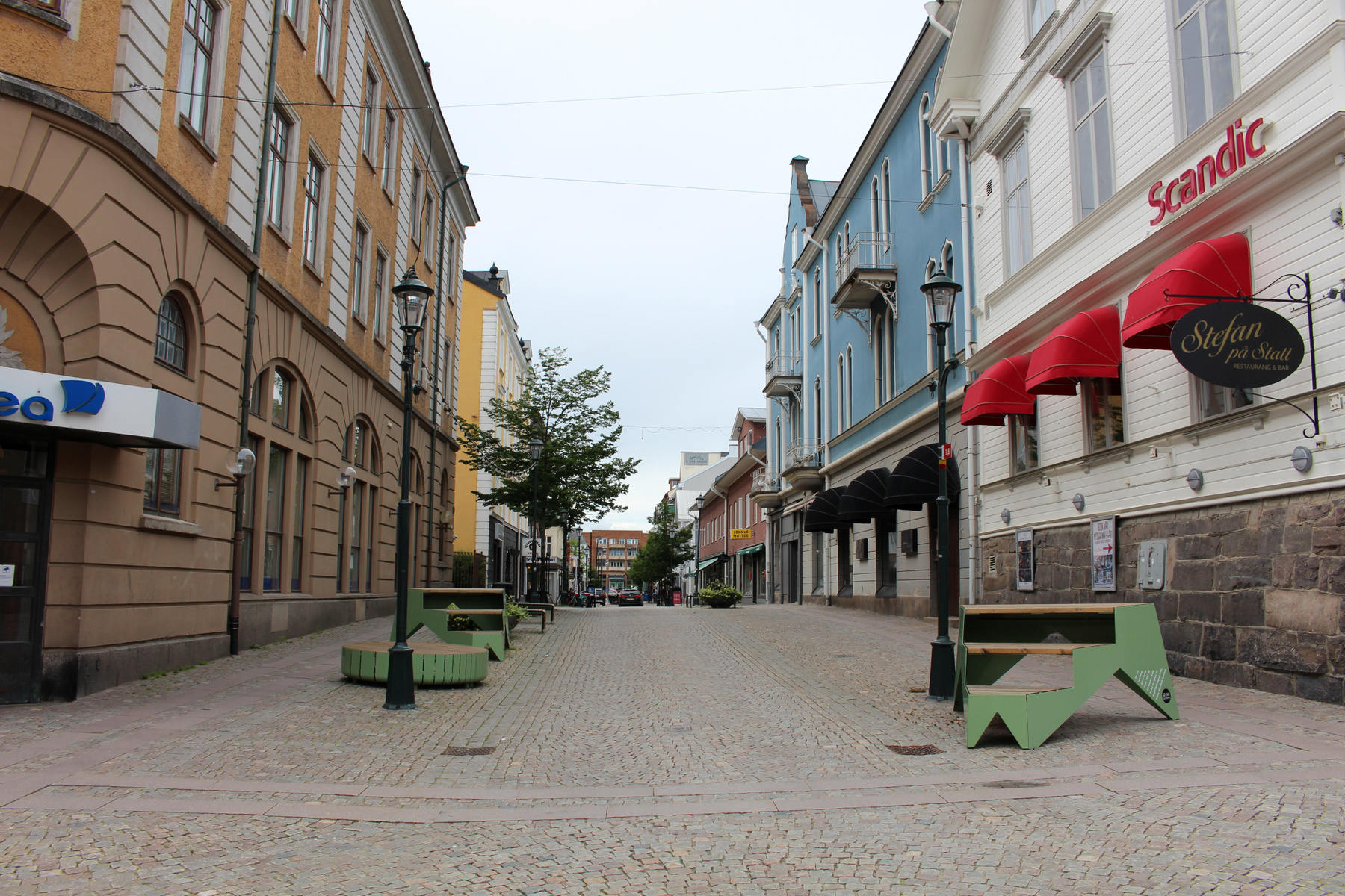 En kullerstensbelagd gata kantad av flervåningsbyggnader i stadsmiljö. På den vänstra sidan syns en gul byggnad med många fönster och en skylt som säger "ea", medan den högra sidan har byggnader i blåa och vita nyanser med "Scandic"-skyltning och röd