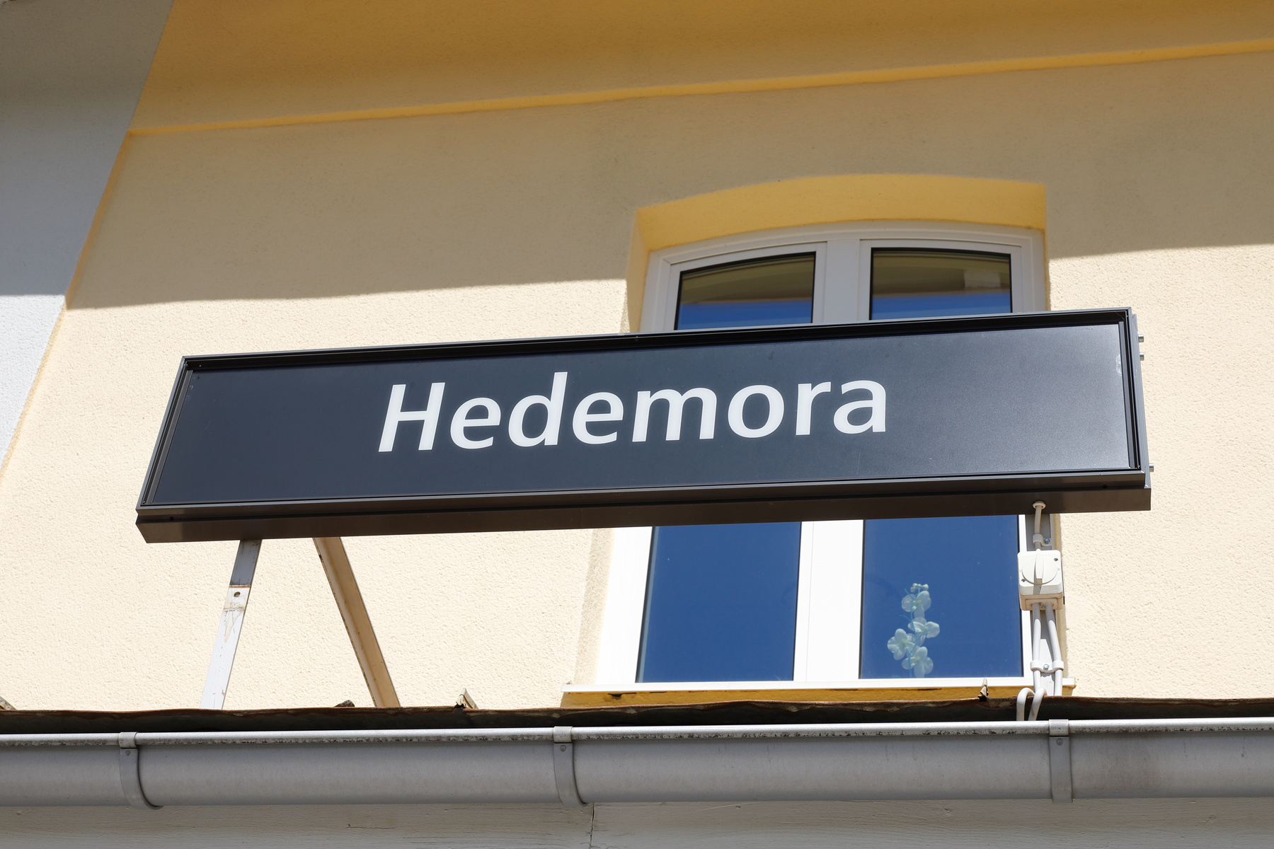 En svart skylt med vit text "Hedemora" är monterad på en ljusgul byggnadsfasad. Under skylten syns ett fönsterparti med blå himmel speglandes i rutorna, samt en takränna nedtill i bilden.