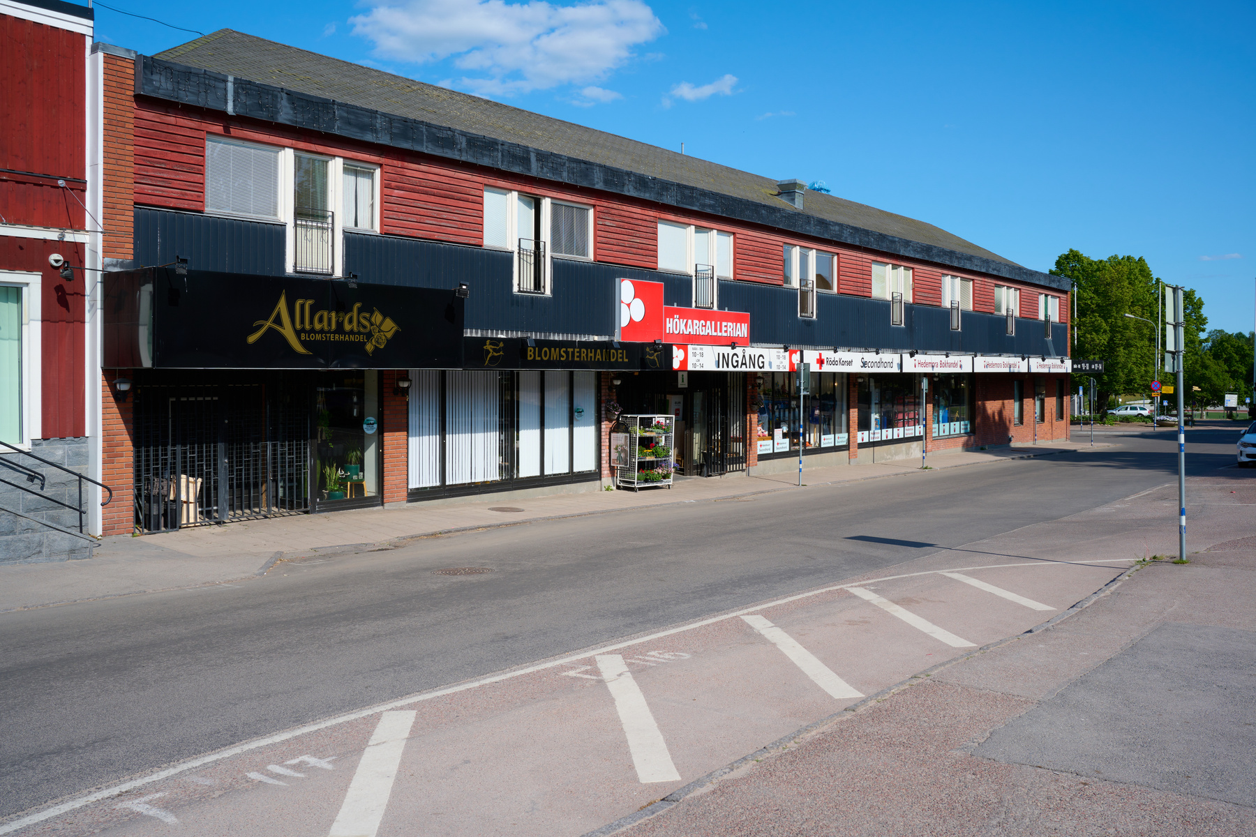 En gathörna med en lång byggnad med röd fasad och svarta detaljer, där flera butiker är belägna på bottenvåningen. På gatan finns en cykelbana och en bil som kör där. Blå himmel med moln ses ovanför byggnaden.