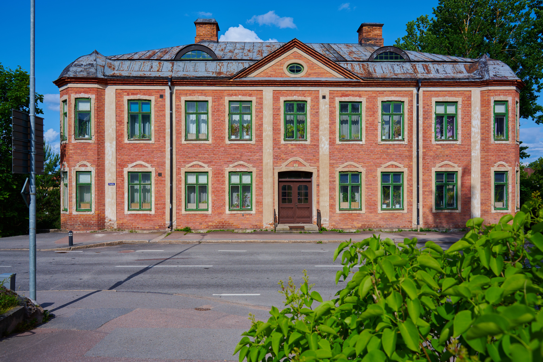 En stor, röd tegelbyggnad med grönmålade fönster står mot en blå himmel med vita moln. Byggnaden har ett grått plåttak med flera kupor och skorstenar, samt en central entré med dubbla bruna dörrar. En asfalterad väg löper framför byggnaden, och en bu