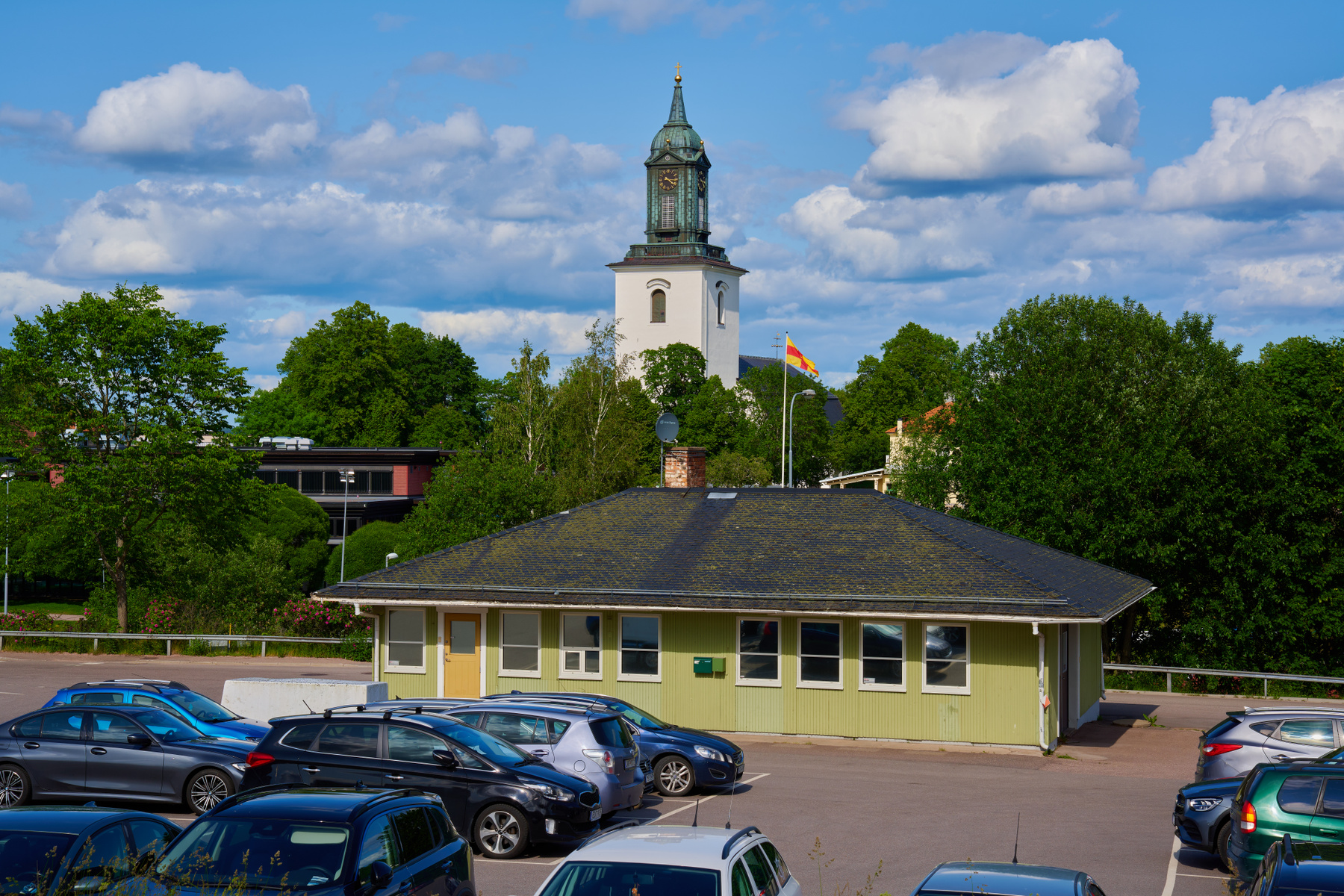 En översiktsbild av en parkeringsplats med flera parkerade bilar framför en enfärgad gulgrön byggnad med ett mörkt tak. I bakgrunden syns frodiga, gröna träd och en kyrka med en vit fasad och ett kopparfärgat kupoltak med klocka. På en flaggstång bre