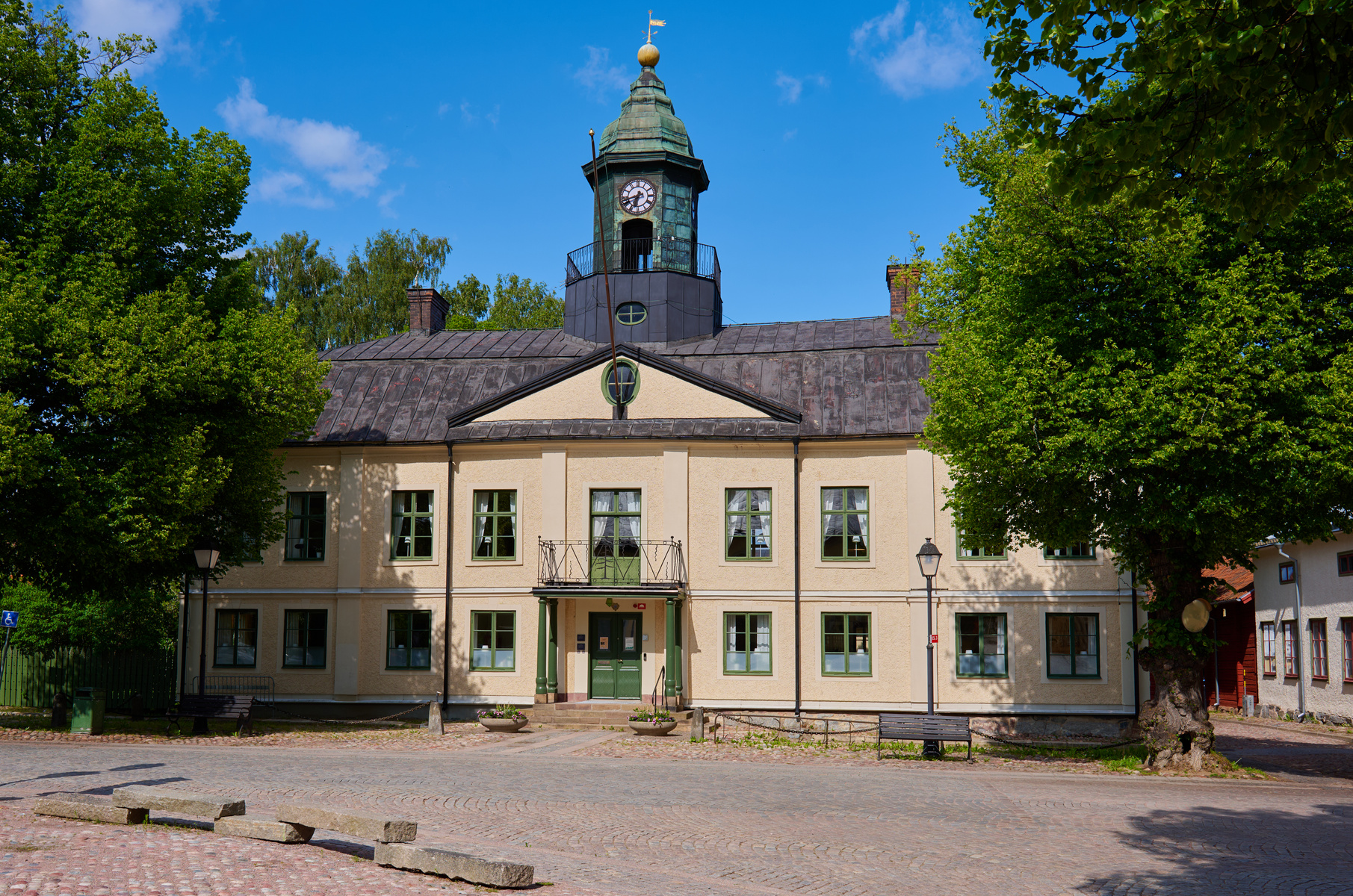 En gul byggnad med grå tak, gröna fönsterluckor och en klocka i tornet syns under en molnig blå himmel. Byggnaden är omgiven av lummiga gröna träd och i förgrunden finns kullerstenar och några liggande stenblock. Till vänster syns även en papperskorg
