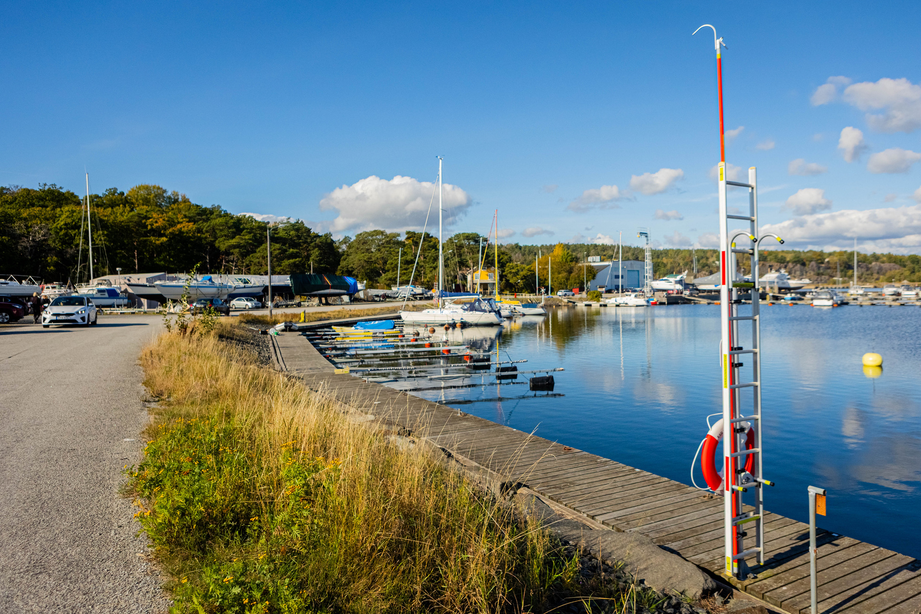 En marin miljö under en klarblå himmel med vita moln. En liten väg går till vänster, med torrlagda båtar och bilar parkerade intill en skogskant. Till höger syns en brygga med förtöjda båtar och en livröddare med en räddningsutrustning fäst vid en st