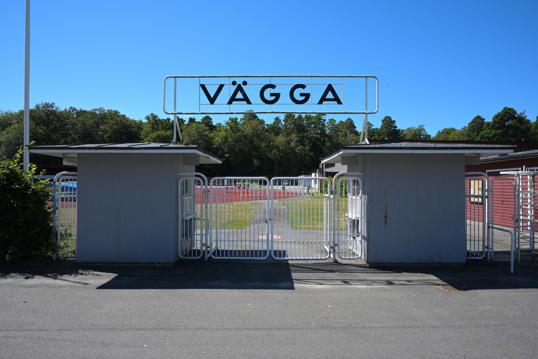 En entré till en arena under en solig dag, med en stor skylt med texten "VÄGGA" över grinden. Två grå byggnader flankerar den stängda vita grinden och bakom syns lövträd och en röd löparbana. Blå himmel utgör bakgrunden.