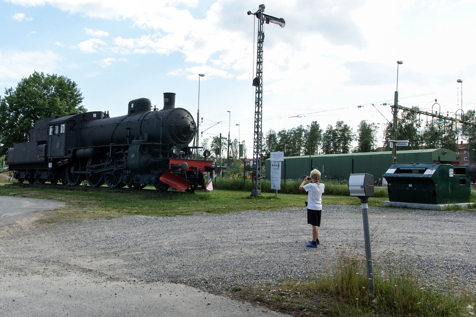 En svart ånglok står på räls bredvid en grusad yta. Ett barn med blont hår fotograferar loket med en mobiltelefon från den grusade ytan. I bakgrunden syns träd, en skylt med texten "PAPPERSFÖRPACKNINGAR" och en grön container. En varningsskylt med te