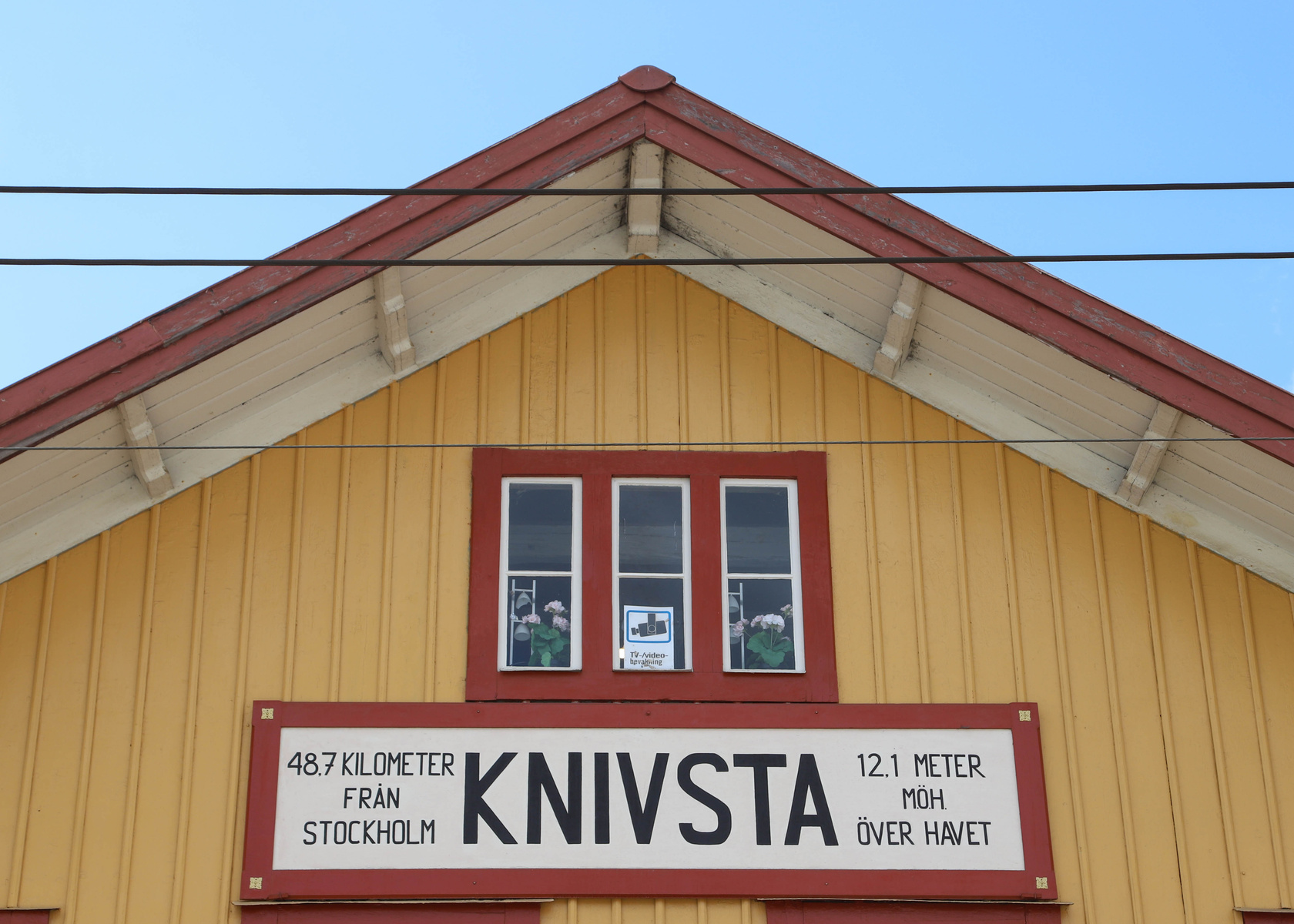 En gul fasad med rött tak och vita detaljer under takfoten. Tre rektangulära fönster sitter centralt med röda fönsterkarmar. En skylt med platsen 'KNIVSTA' samt information om avstånd till Stockholm och höjd över havet. Flera elledningar korsar bilde