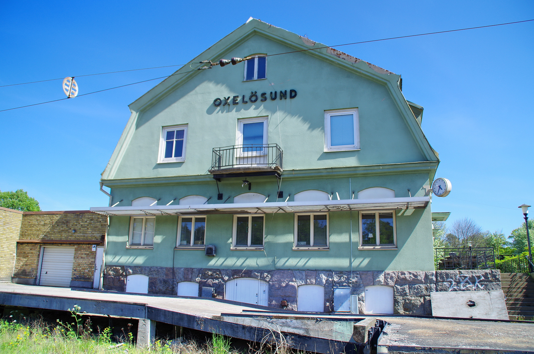 En ljusgrön byggnad med "OXELÖSUND" skrivet på fasaden står under en klarblå himmel. Byggnaden har flera fönster, en balkong och en yta av natursten i botten. Bredvid står en tegelbyggnad med garageport och en klocka är monterad på den ljusgröna bygg