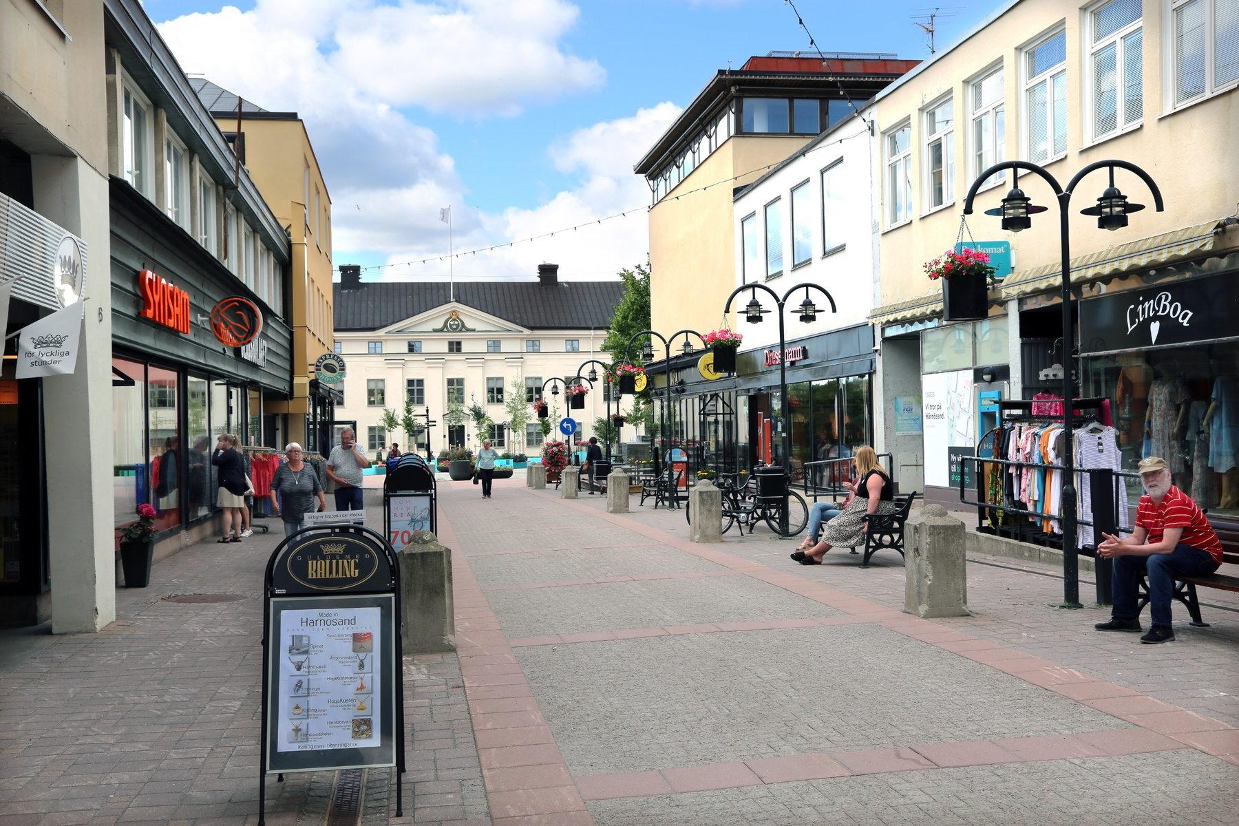 En gata i en stad med byggnader på båda sidor. Personer syns gående och sittande på bänkar. Det finns skyltar, blommor och butiksfönster. En stor byggnad i bakgrunden är synlig under en blå himmel med moln.