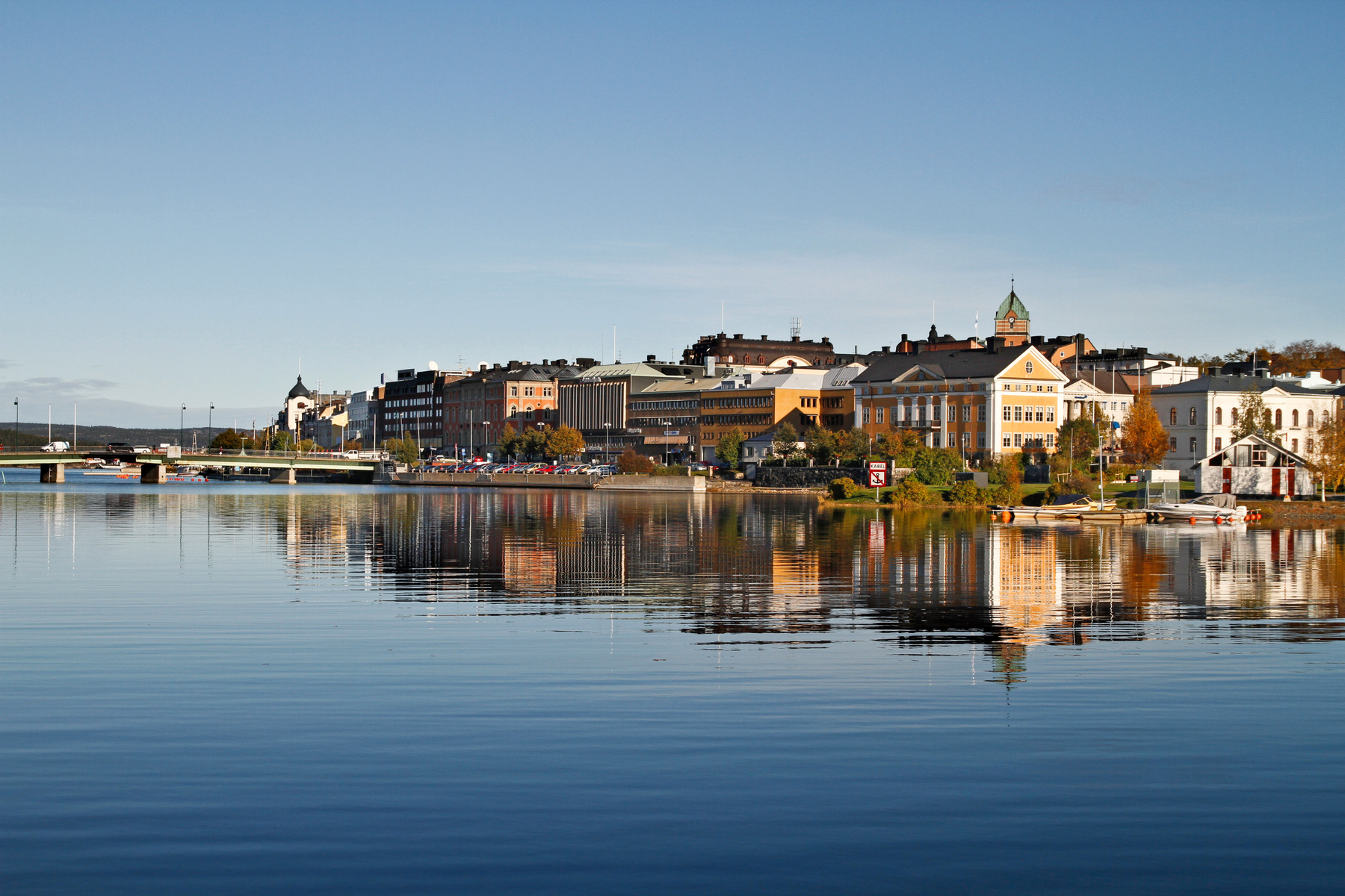 En panoramabild över en stad vid vattnet, med byggnader som speglar sig i vattnet. I förgrunden syns vattenytan och i bakgrunden en stadssiluett med olika byggnader och, till vänster, en bro över vattnet med bilar på.
