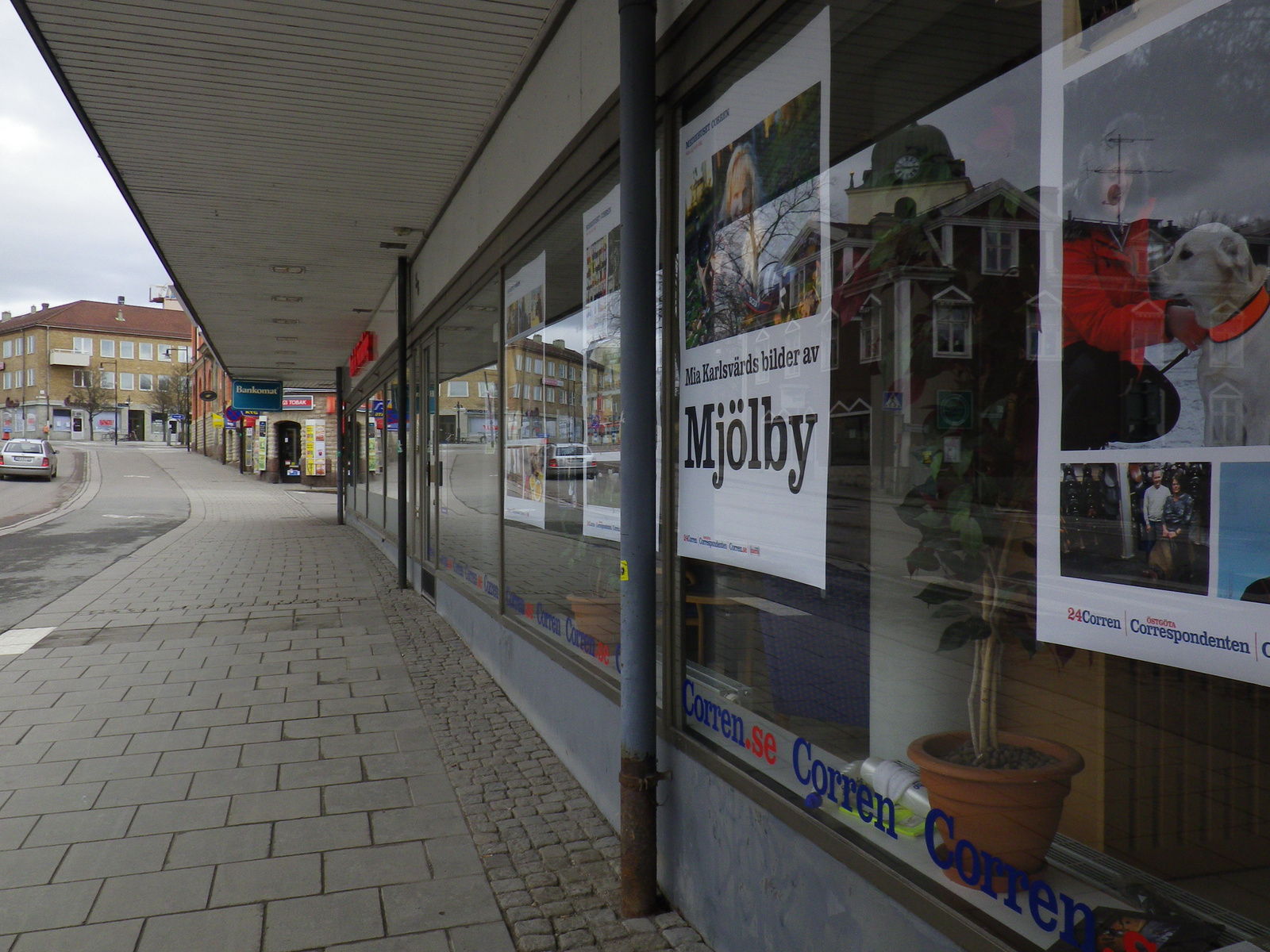 Bilden visar en gata med en trottoar i förgrunden och byggnader längs hörnet. På höger sida finns en fasad med stora skyltfönster, där affischer med texten "Mia Karlsvärds bilder av Mjölby" och "Corren.se Corren" syns. Ett brunt lerkärl med en växt r
