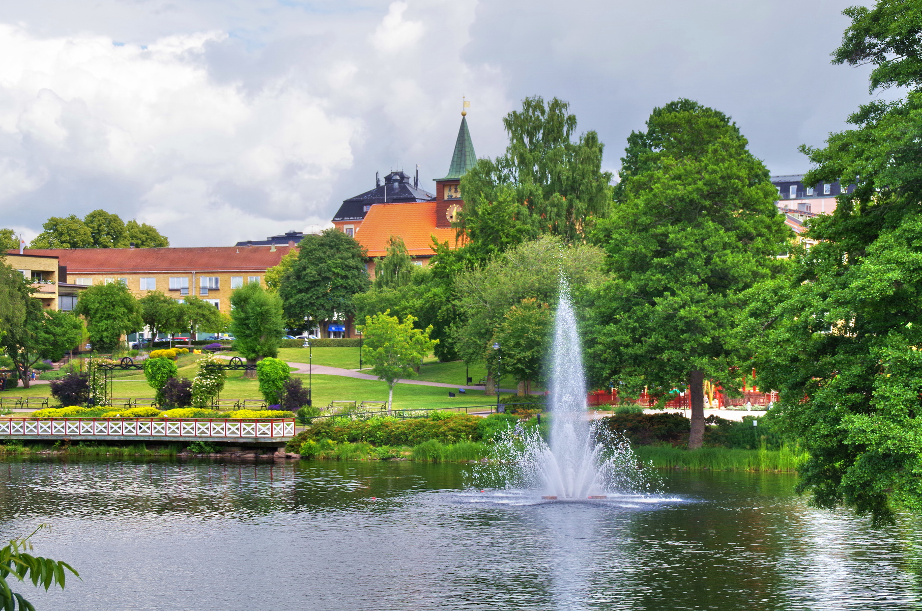 En bild visar en park med en sjö i förgrunden, där en fontän sprutar vatten. I bakgrunden syns flera träd, grön gräsmatta, en gångväg, samt byggnader med röda och gula fasader. Molnig himmel syns ovanför.