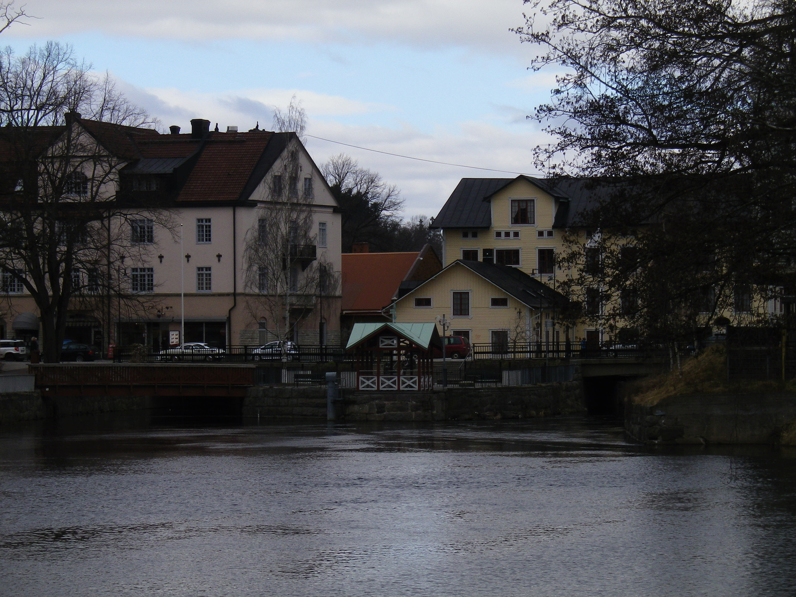 En bild som visar en flod eller å med hus på båda sidor av vattnet. Flera broar korsar vattnet. Träd med kala grenar står längs stranden och framför byggnaderna. Himlen är delvis mulen med ljusblå fläckar. Flera fordon syns bland husen.
